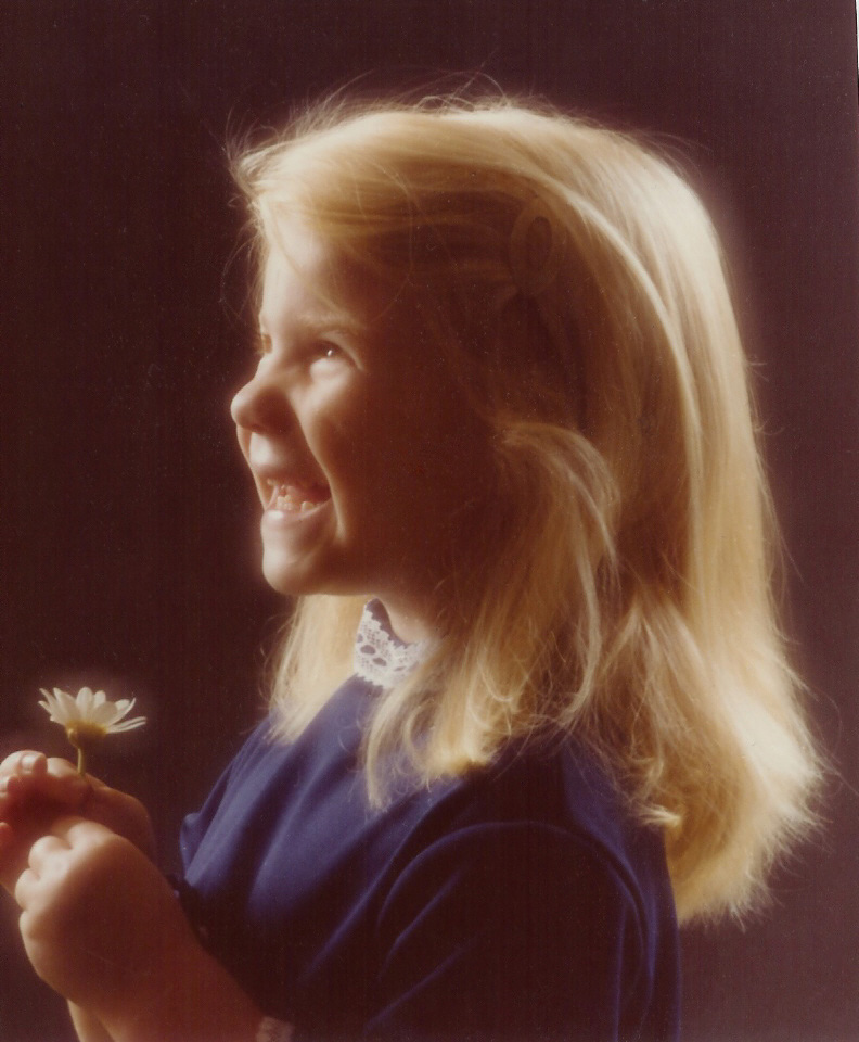 young girl in a blue dress with long blonde hair holding a daisy smiling